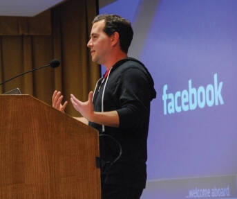 A man speaking at a podium in front of a screen displaying the Facebook logo.